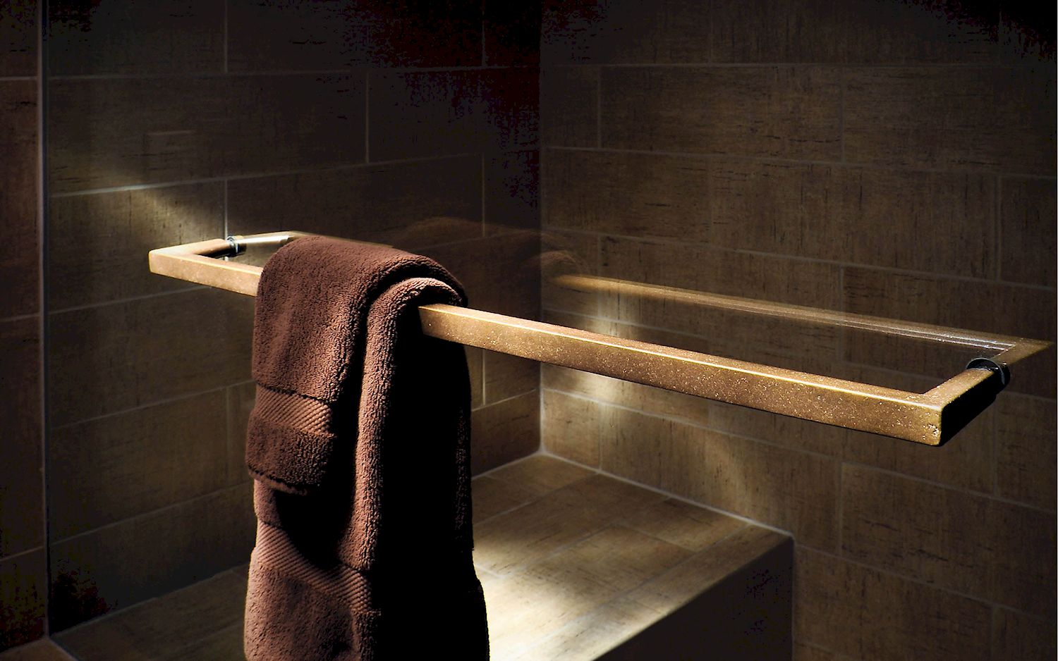 a brown towel hangs on a bronze towel rack mounted to a glass shower door. The shower is tiled with a brown tile and a bench is visible inside. 