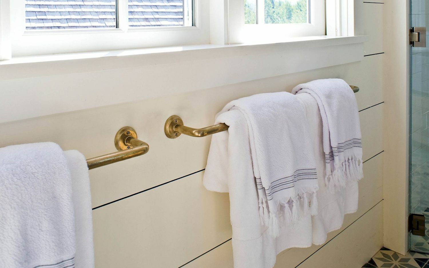 two silicon bronze towel bars are mounted to a white wall beneath a window. Two layers of luxurious white towels hand on the bars.